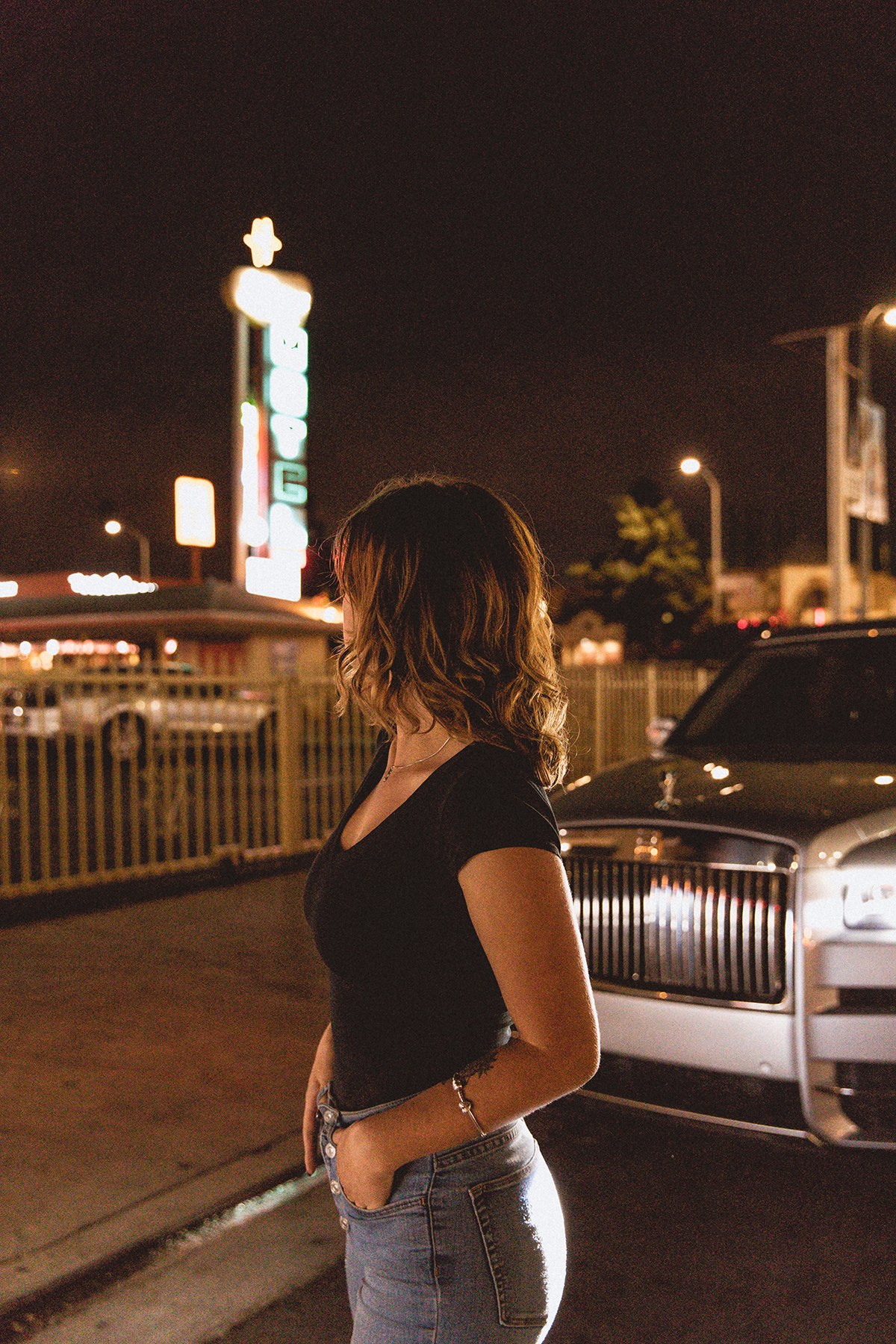 Rolls-Royce Cullinan – woman from behind beside the car, under a vintage motel neon sign in Los Angeles at night, photo Jon Filin